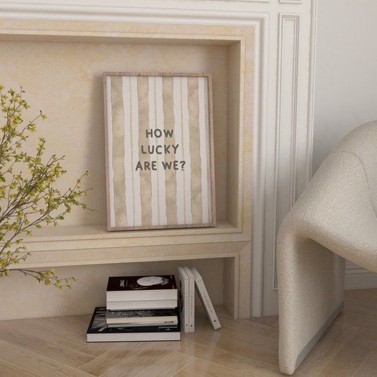 Framed picture with 'How Lucky Are We?' text on a wall with books and a plant below.