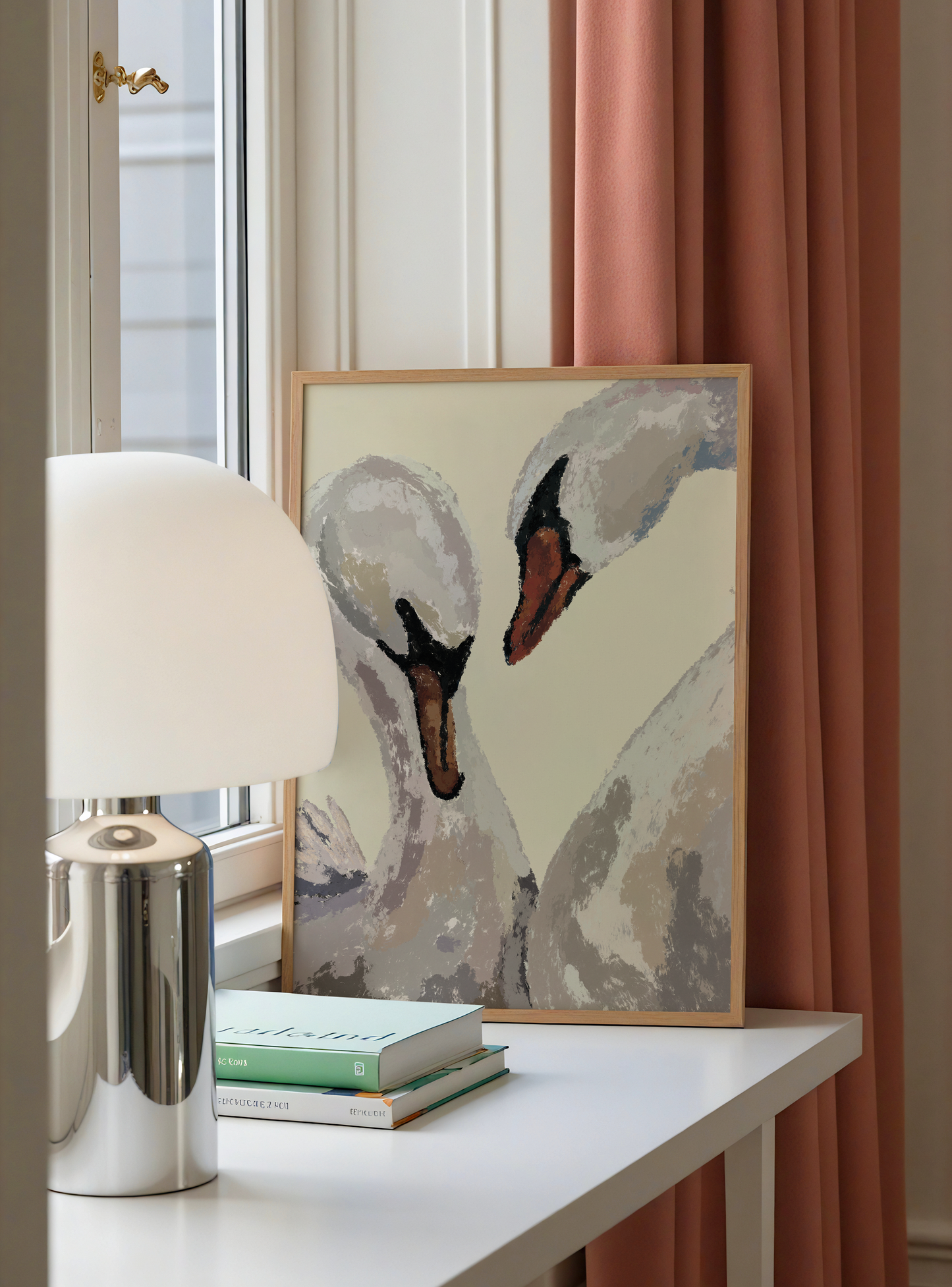 A framed painting of two swans leaning against a white table in a styled room setting, next to a modern silver and white table lamp and stacked books, with a dusty rose curtain in the background.