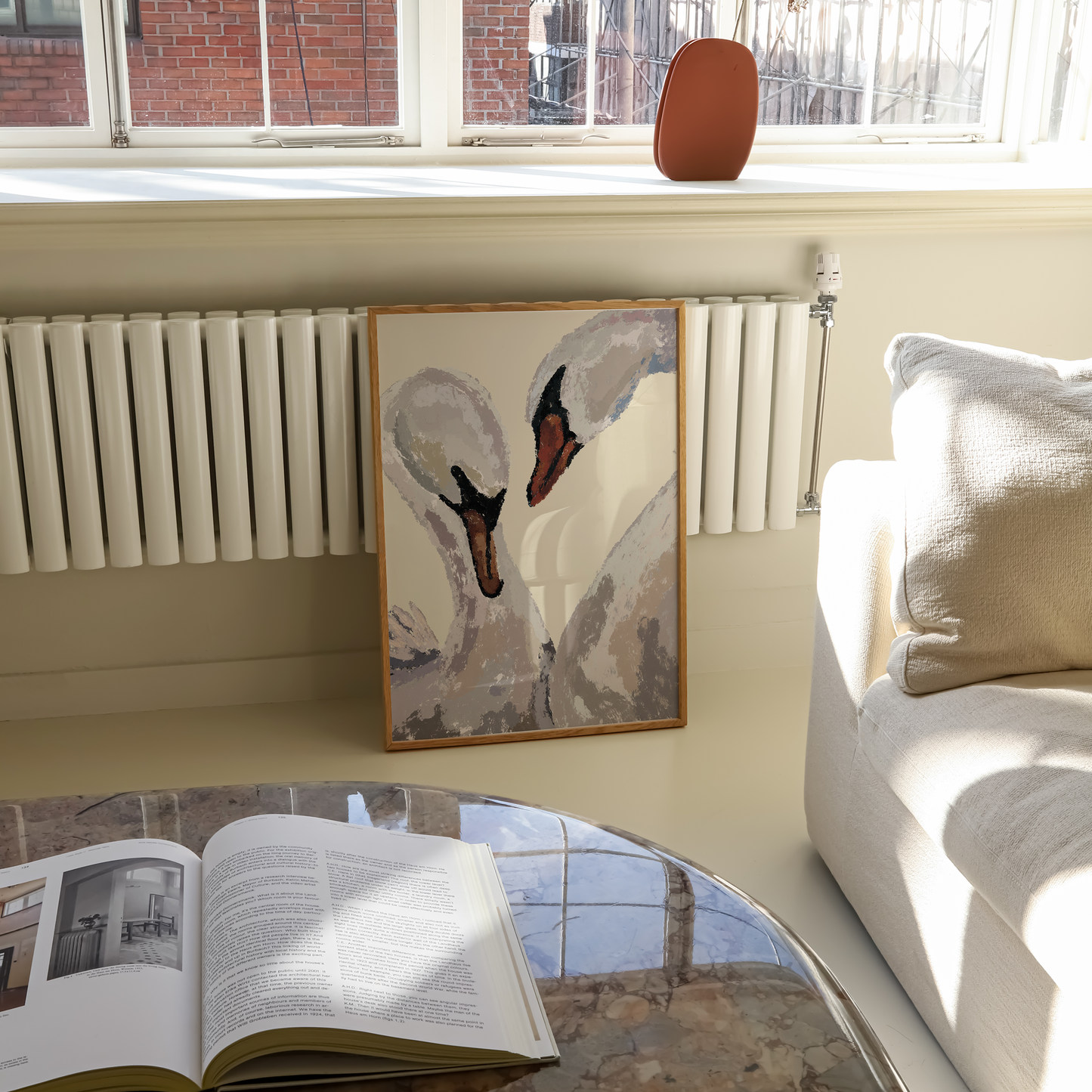 A framed painting of two swans leaning against a cream-colored wall in a sunlit living room, with a white radiator below, a sofa to the right, and an open book on a marble coffee table in the foreground.