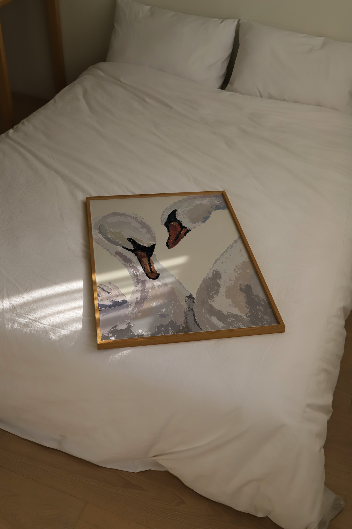 A framed art print depicting two swans, displayed on a neatly made white bed with pillows. The print features a light wood frame and is photographed in a home setting with natural light casting shadows on the duvet.