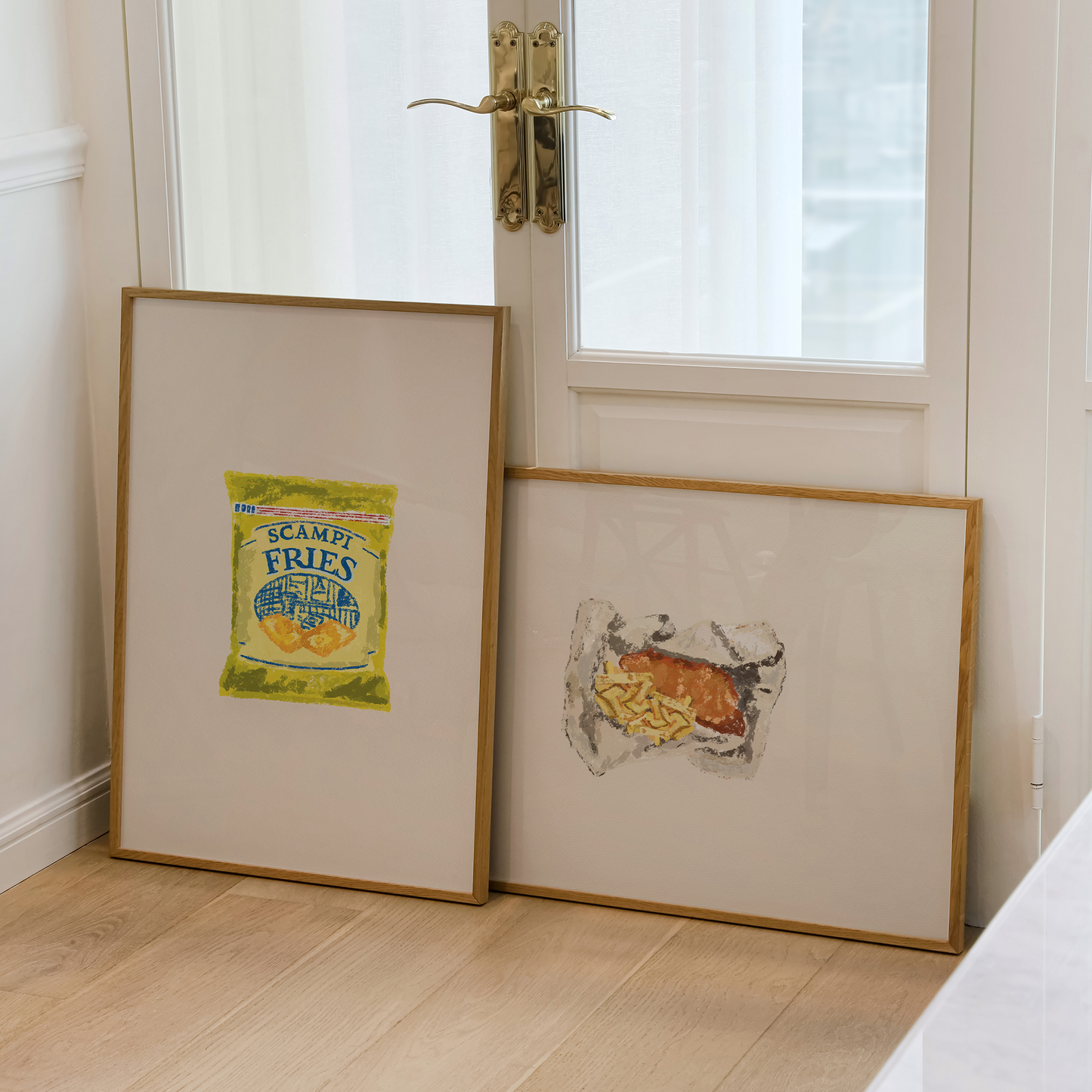 Two framed art prints leaning against a white door in a bright room with light wood flooring. The left print features a vintage-style 'Sesame Fries' illustration, and the right print shows a whimsical cow drawing.