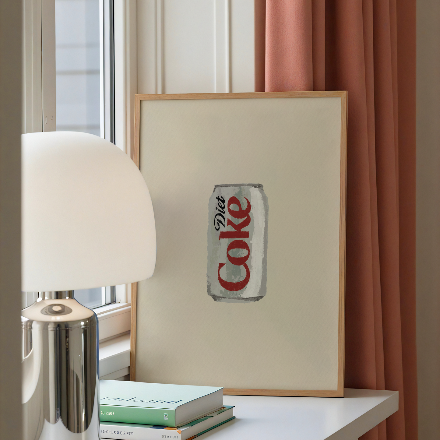 A framed art print featuring a vintage-style painted Coca-Cola can on an off-white background, leaning on a white table next to a silver lamp and books, with a window and peach curtains in the background.