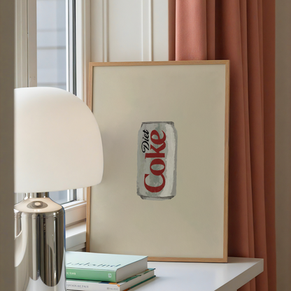 A framed art print featuring a vintage-style painted Coca-Cola can on an off-white background, leaning on a white table next to a silver lamp and books, with a window and peach curtains in the background.