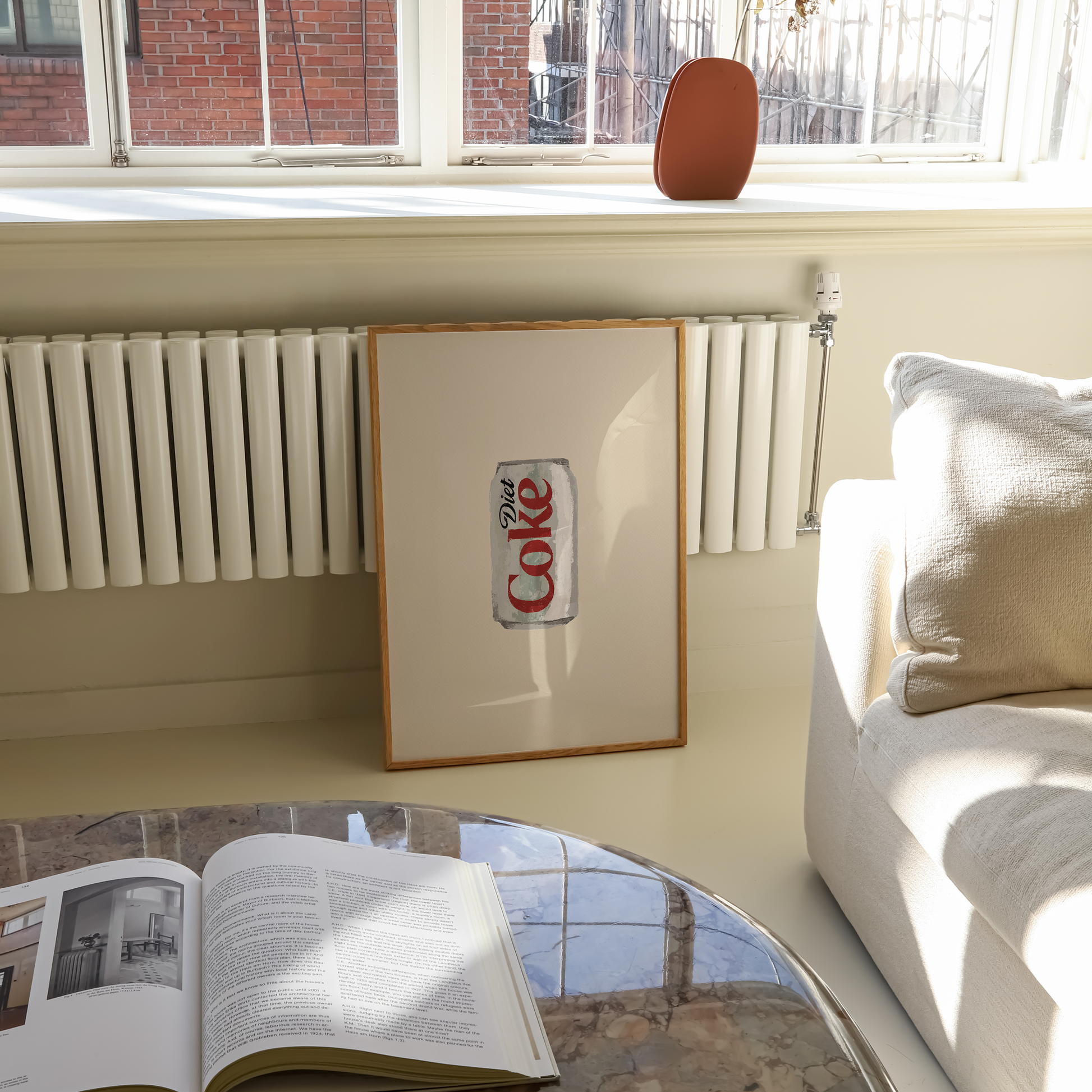 A framed print featuring the vintage Coca-Cola logo leaning against a cream wall in a bright living room. Sunlight streams in from a window behind, showing a radiator and brick building outside, with a sofa and a marble coffee table in the foreground.