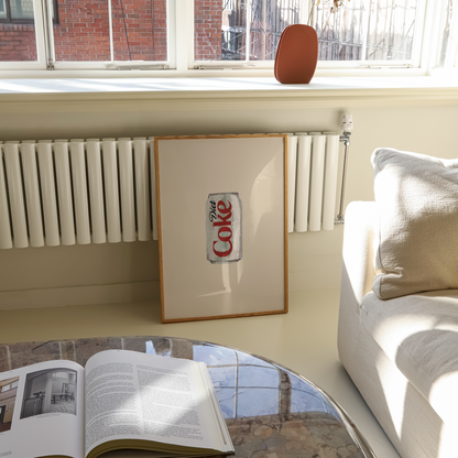 A framed print featuring the vintage Coca-Cola logo leaning against a cream wall in a bright living room. Sunlight streams in from a window behind, showing a radiator and brick building outside, with a sofa and a marble coffee table in the foreground.