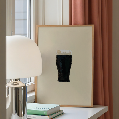 A framed minimalist art print featuring a black pint of beer with white foam on a cream background, styled on a white table next to a silver lamp and books, with a dusty rose curtain in the background.