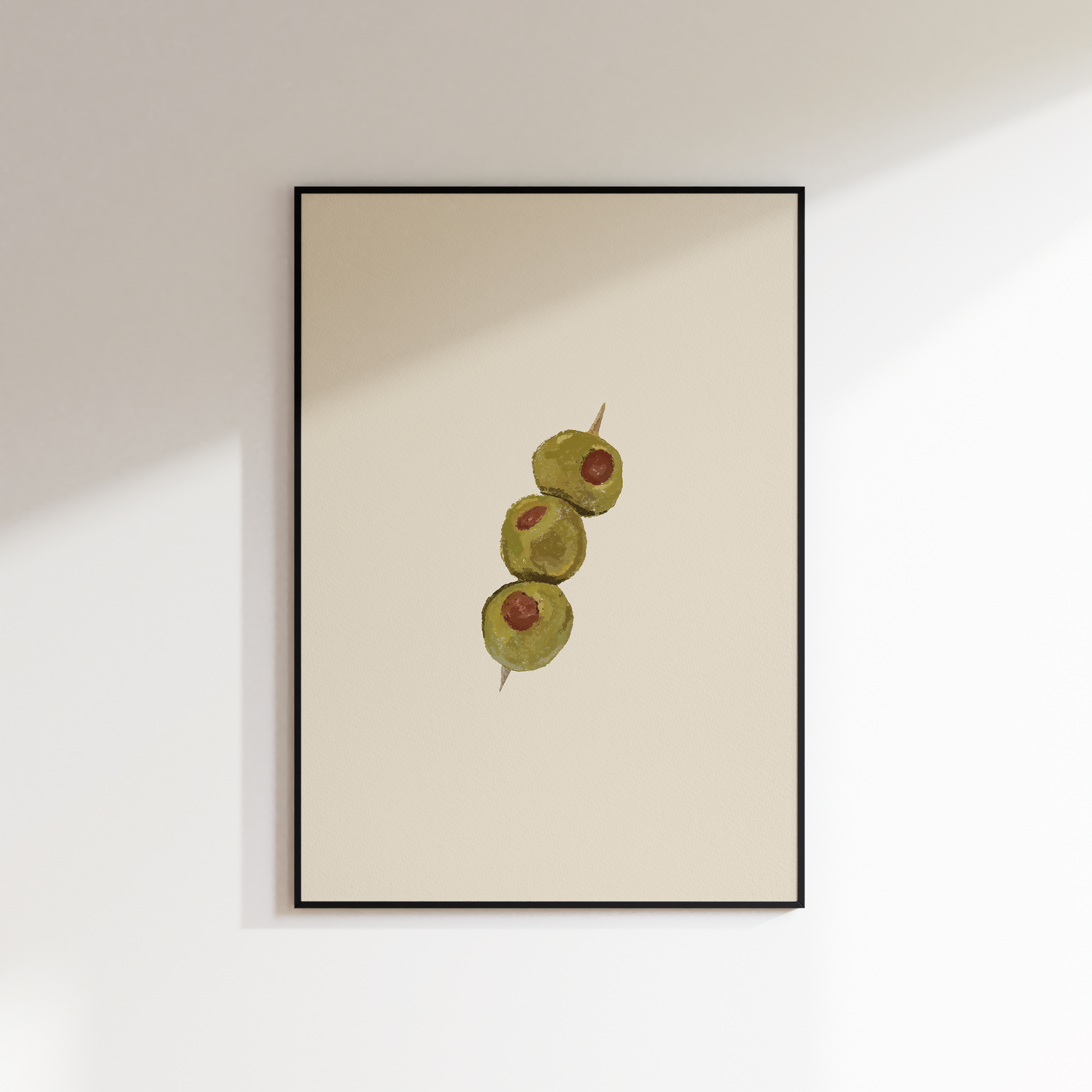 A minimalist framed art print featuring three green olives on a warm cream background, hung on a white wall with soft diagonal shadows.