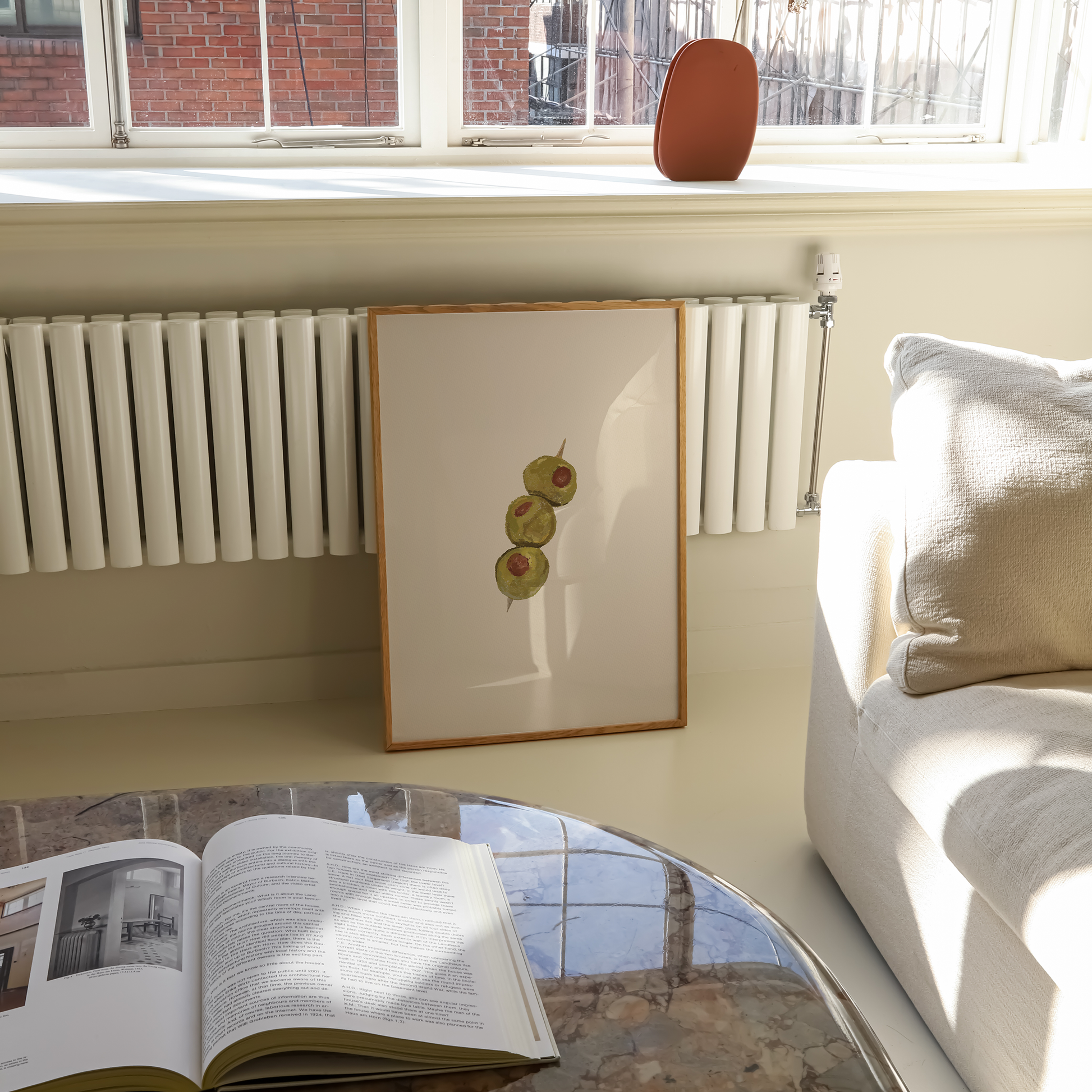 A framed art print featuring three green olives with red pimentos on a cream background, leaning against a white radiator in a bright living room next to a white sofa.