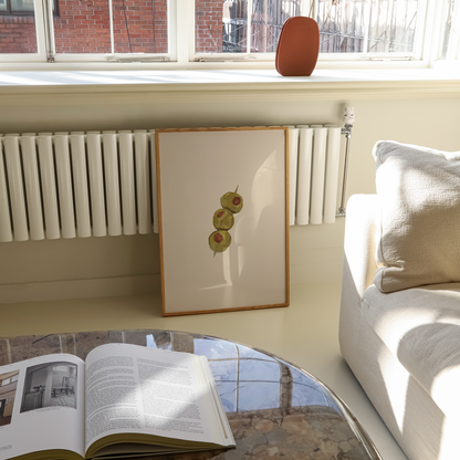 A framed art print featuring three green olives with red pimentos on a cream background, leaning against a white radiator in a bright living room next to a white sofa.