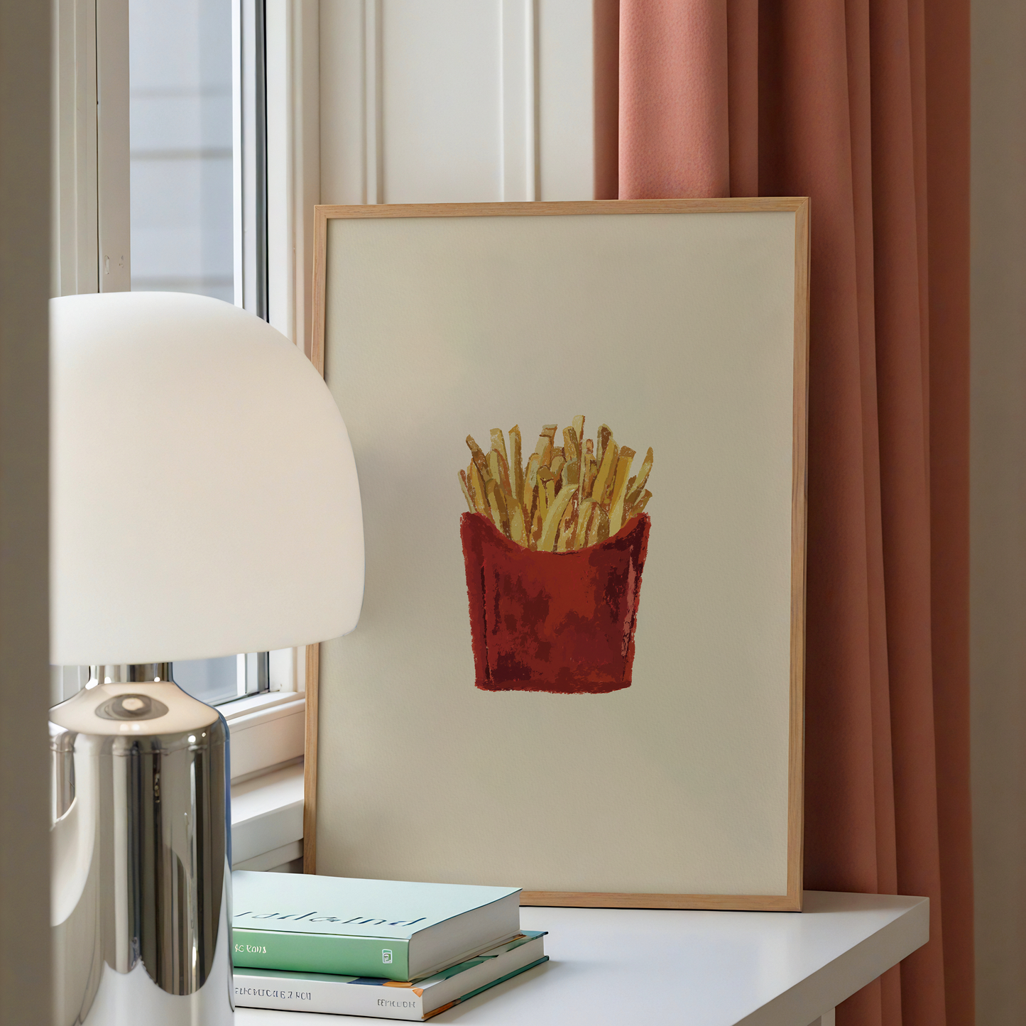A framed art print depicting a red paper bag of french fries on an off-white background, styled on a white table next to a silver lamp and books, with a pink curtain in the background.