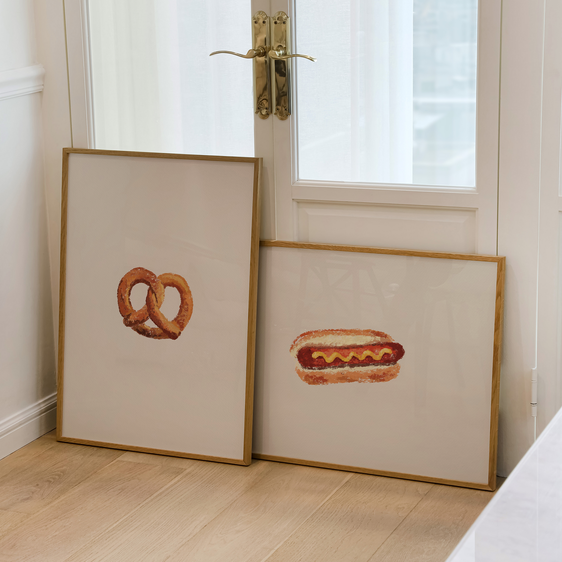 Two framed art prints leaning against a white door on a light wood floor. One print shows a pretzel, and the other shows a hot dog, both in simple gold frames.