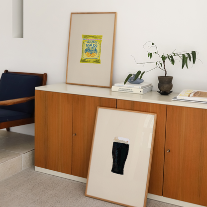 A styled interior scene showing two framed art prints leaning and standing against a white wall above a wooden credenza. The standing print has a yellow-green graphic, and the leaning print features a black abstract shape on a cream background.